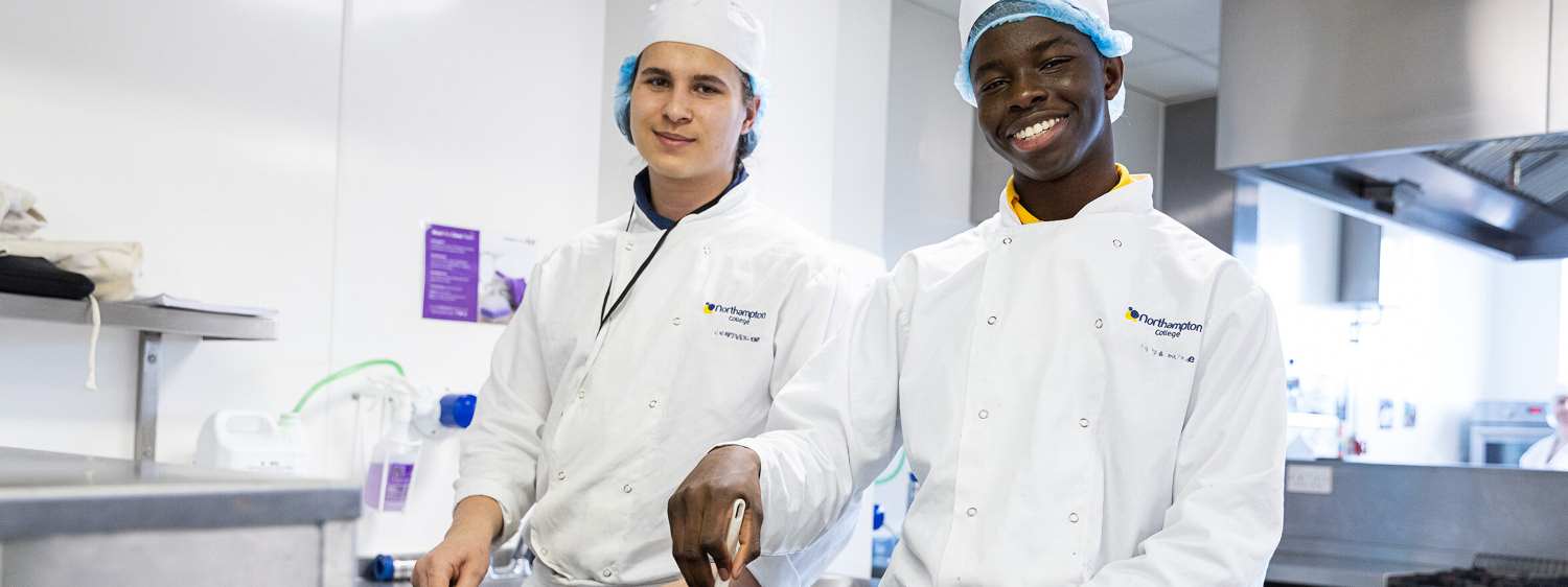 Two male student posse in the College kitchen wearing chef whites.