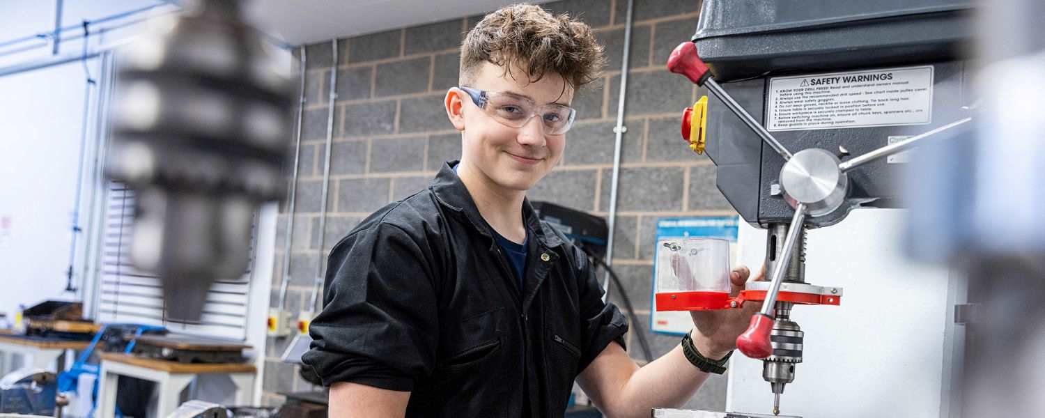 Image shows young student in a workshop, using equipment and smiling to camera.