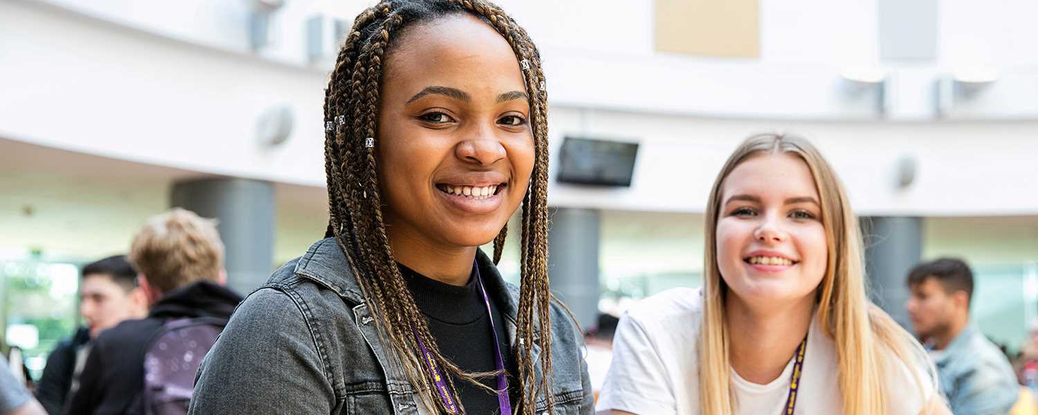 Two female students sitting in the canteen and smiling to camera.